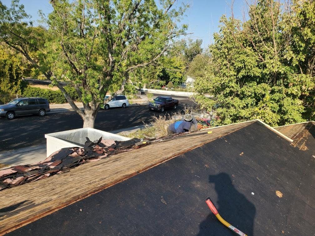 Roof mid tear-off with old shingles piled at eave, bare decking, felt underlayment and a worker clearing debris, in West Jordan, UT