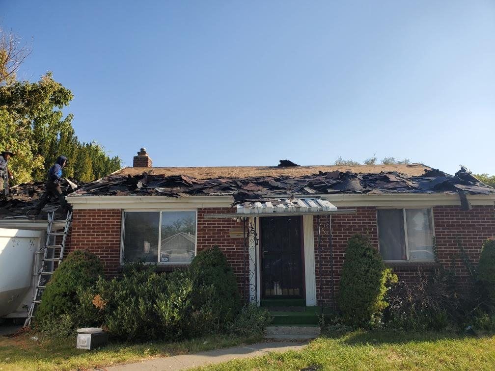 Brick ranch house during roof tear-off, torn shingles over bare decking, workers and ladder at left