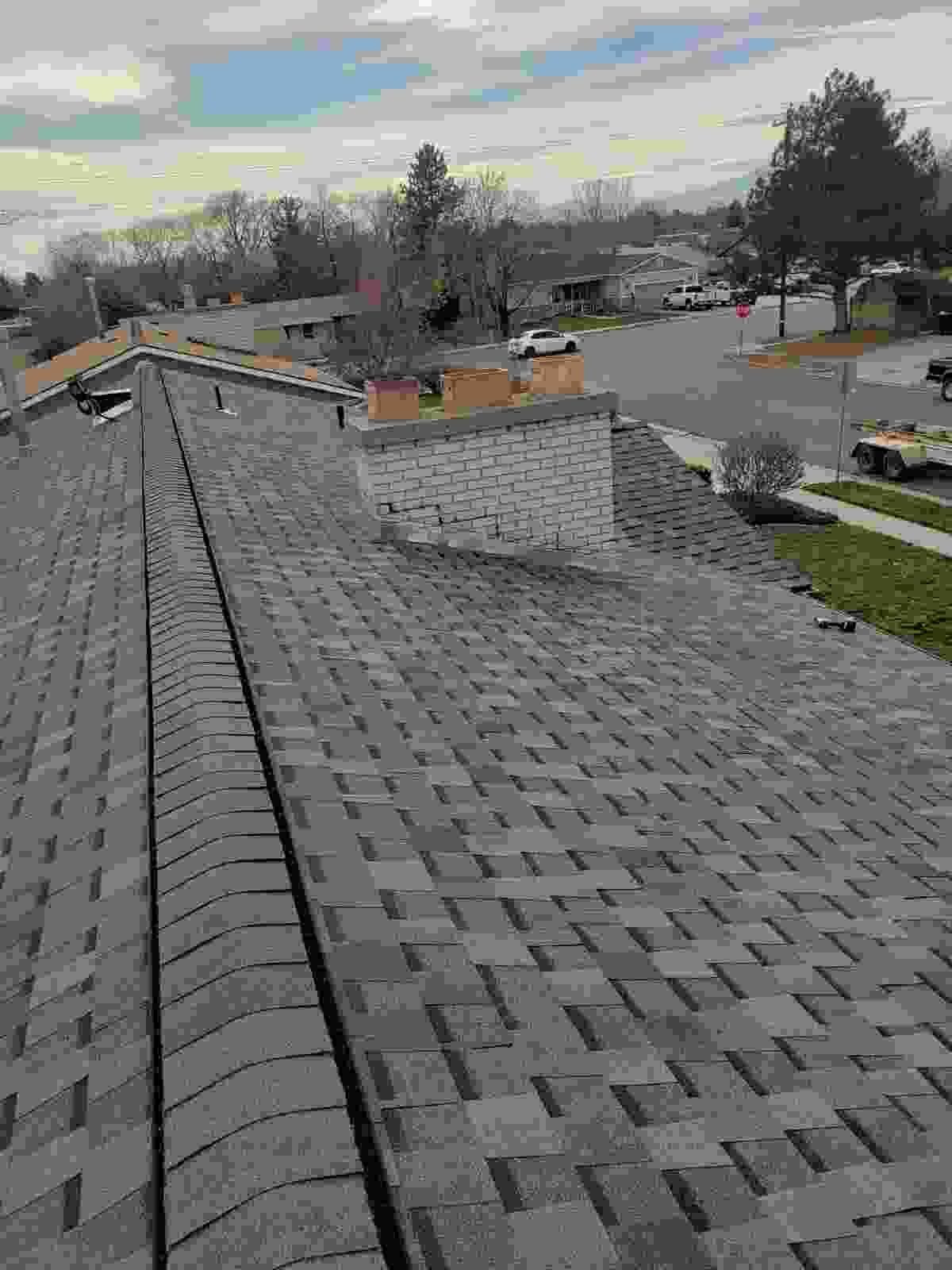 Gray asphalt shingle roof with ridge cap and light masonry chimney, suburban street and bare trees beyond, in Murray, UT