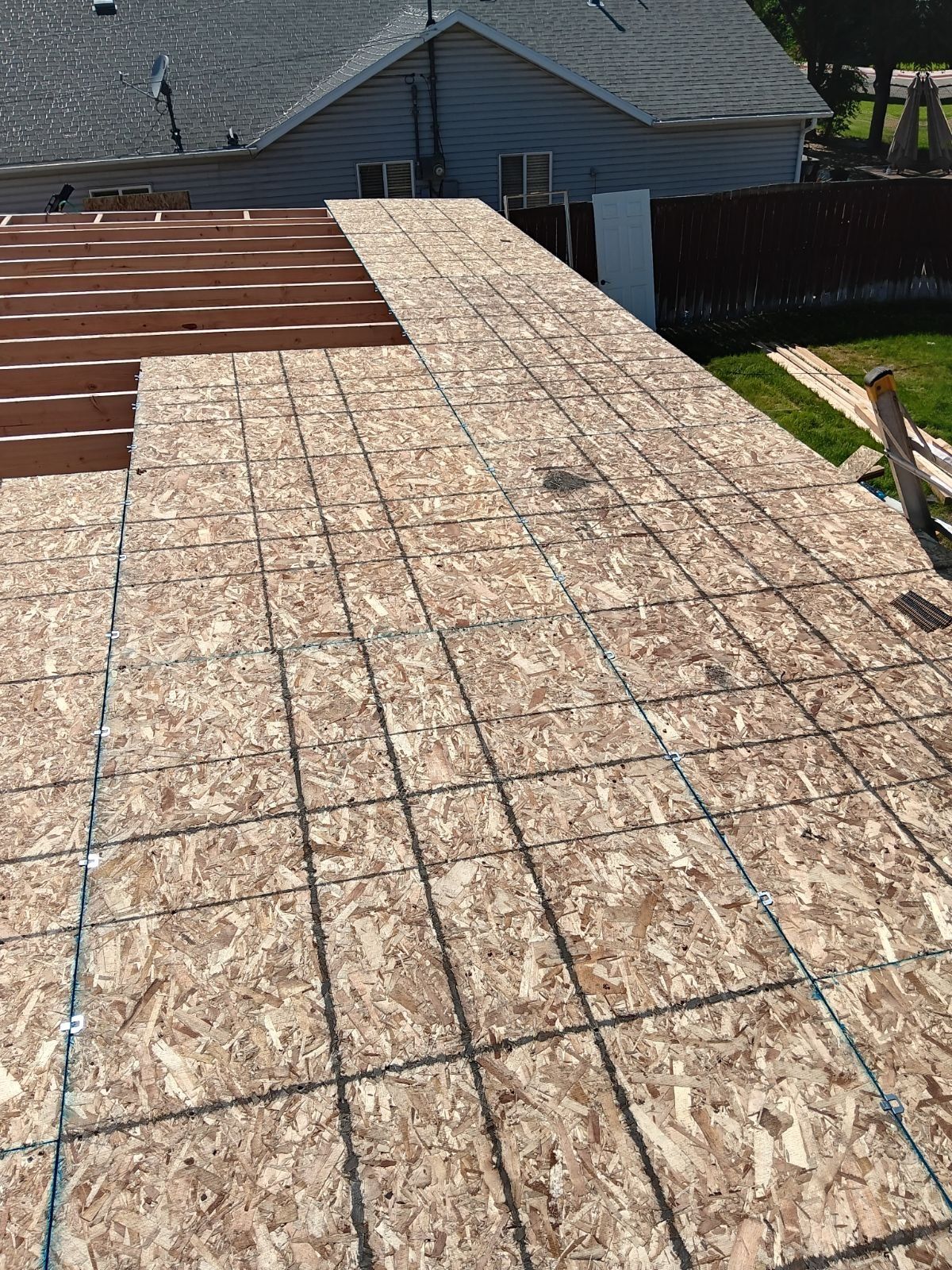 Bare OSB roof decking with chalk lines, exposed rafters at left, house and wood fence behind