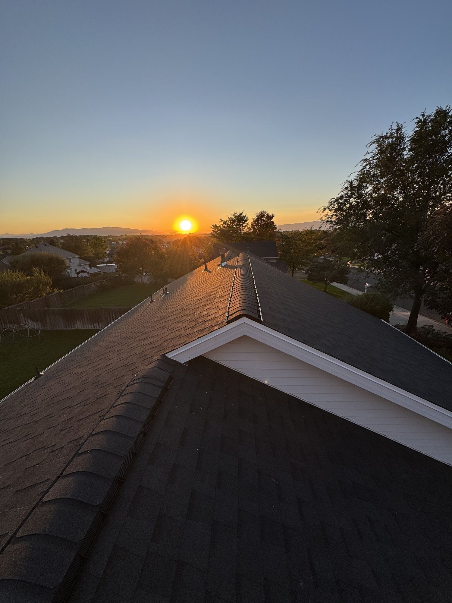 Finished dark asphalt shingle roof ridge at sunset with pipe vents, ridge cap and white gable trim
