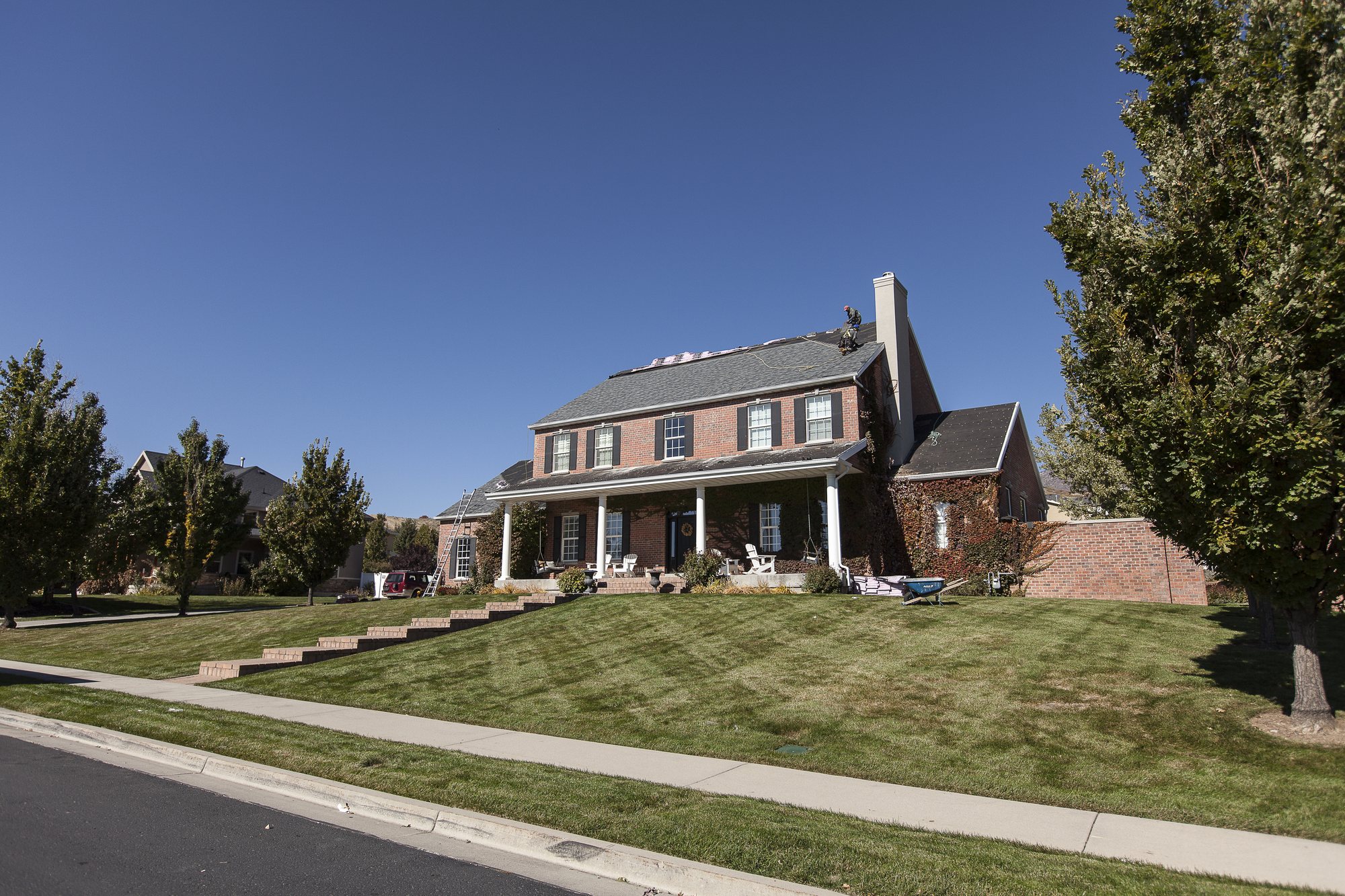 Workers on the steep asphalt shingle roof of a two-story brick house, ladder and materials below, in Clinton, UT