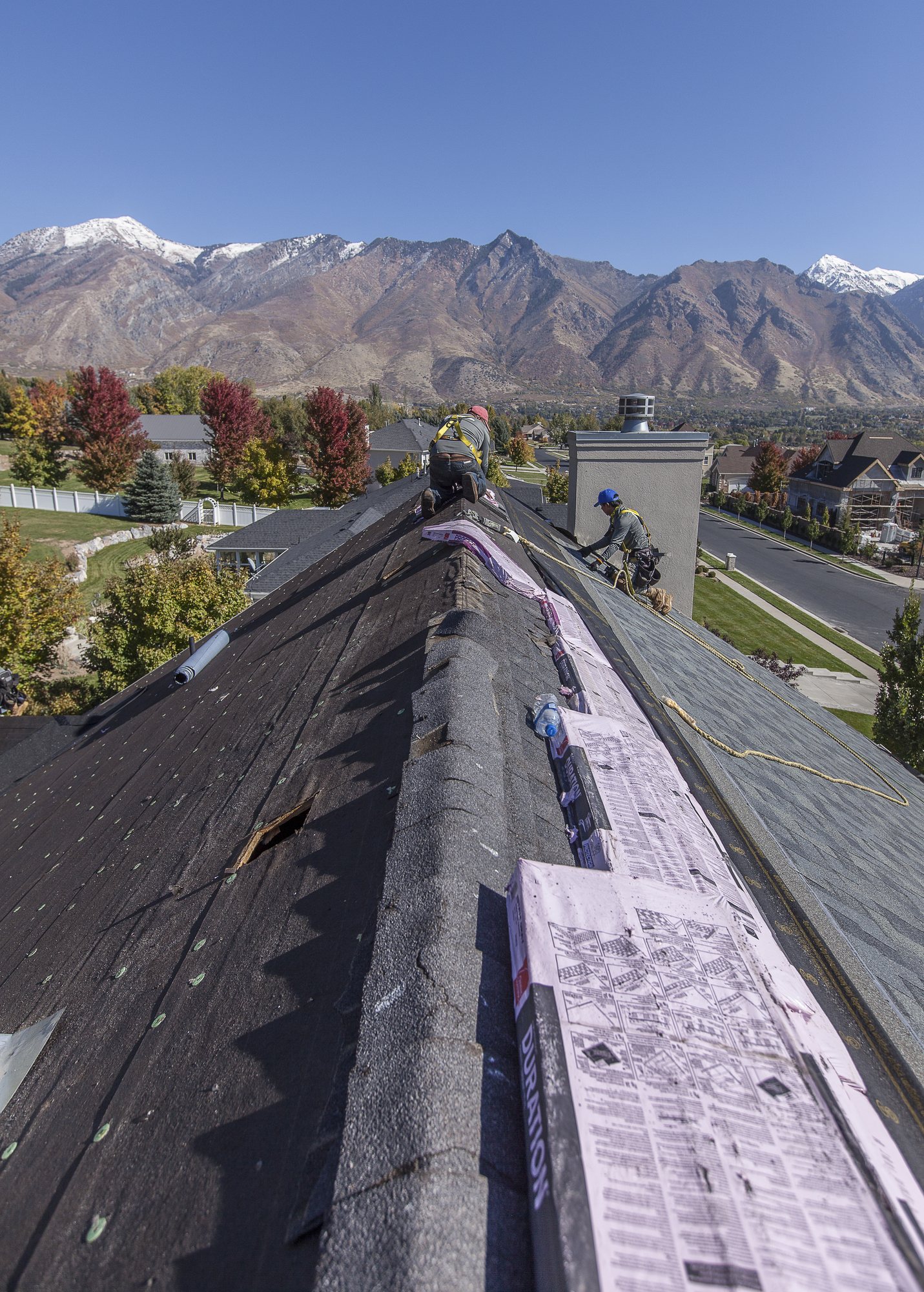 Two harnessed roofers at a stripped shingle ridge with new bundles staged, snow-topped mountains in the background, in Kearns, UT