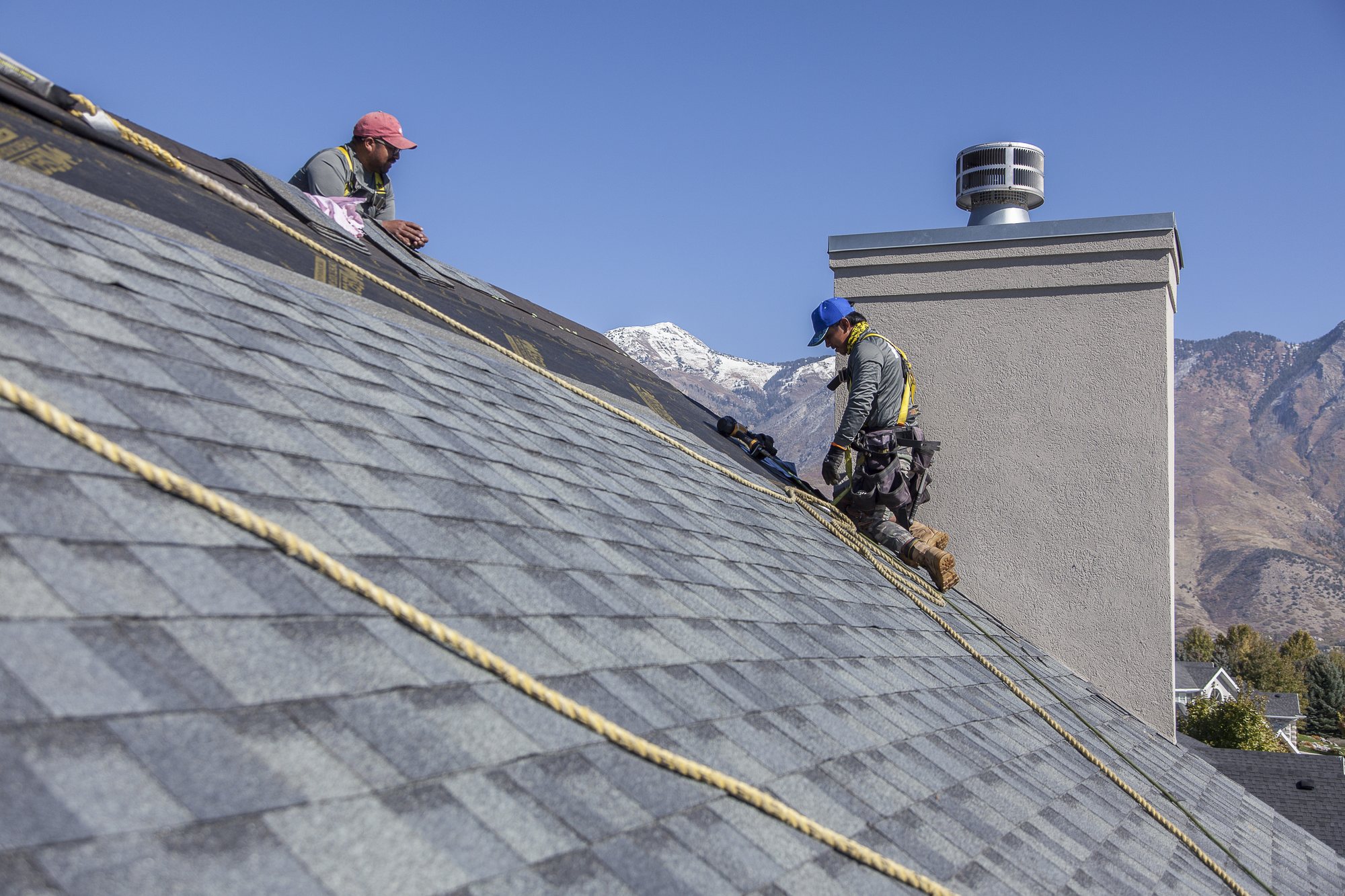Two harnessed roofers laying gray asphalt shingles on a steep roof near a stucco chimney, snowy peaks behind