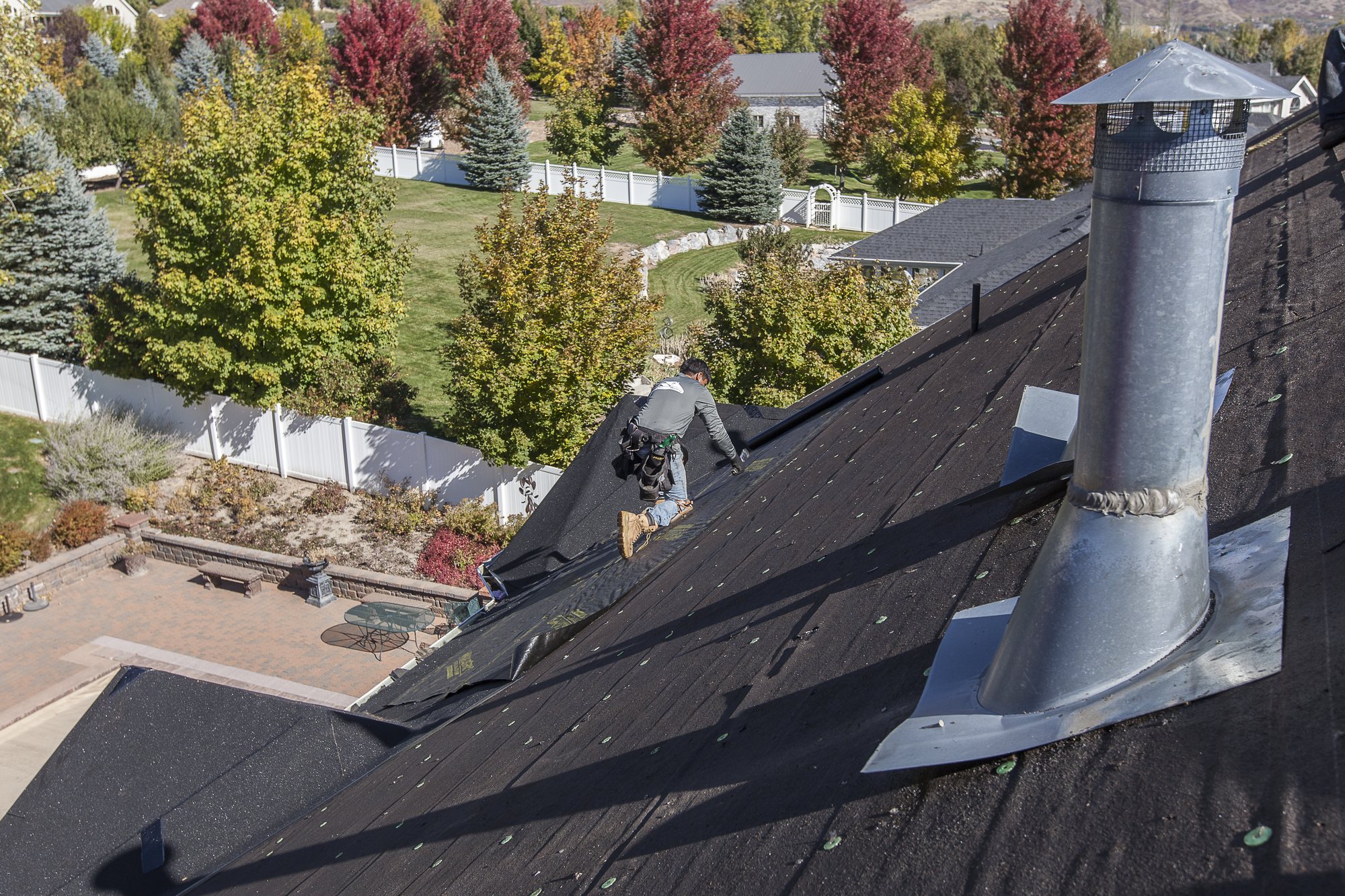 Roofer kneeling on steep roof covered in black underlayment next to a metal flue pipe with sheet-metal flashing