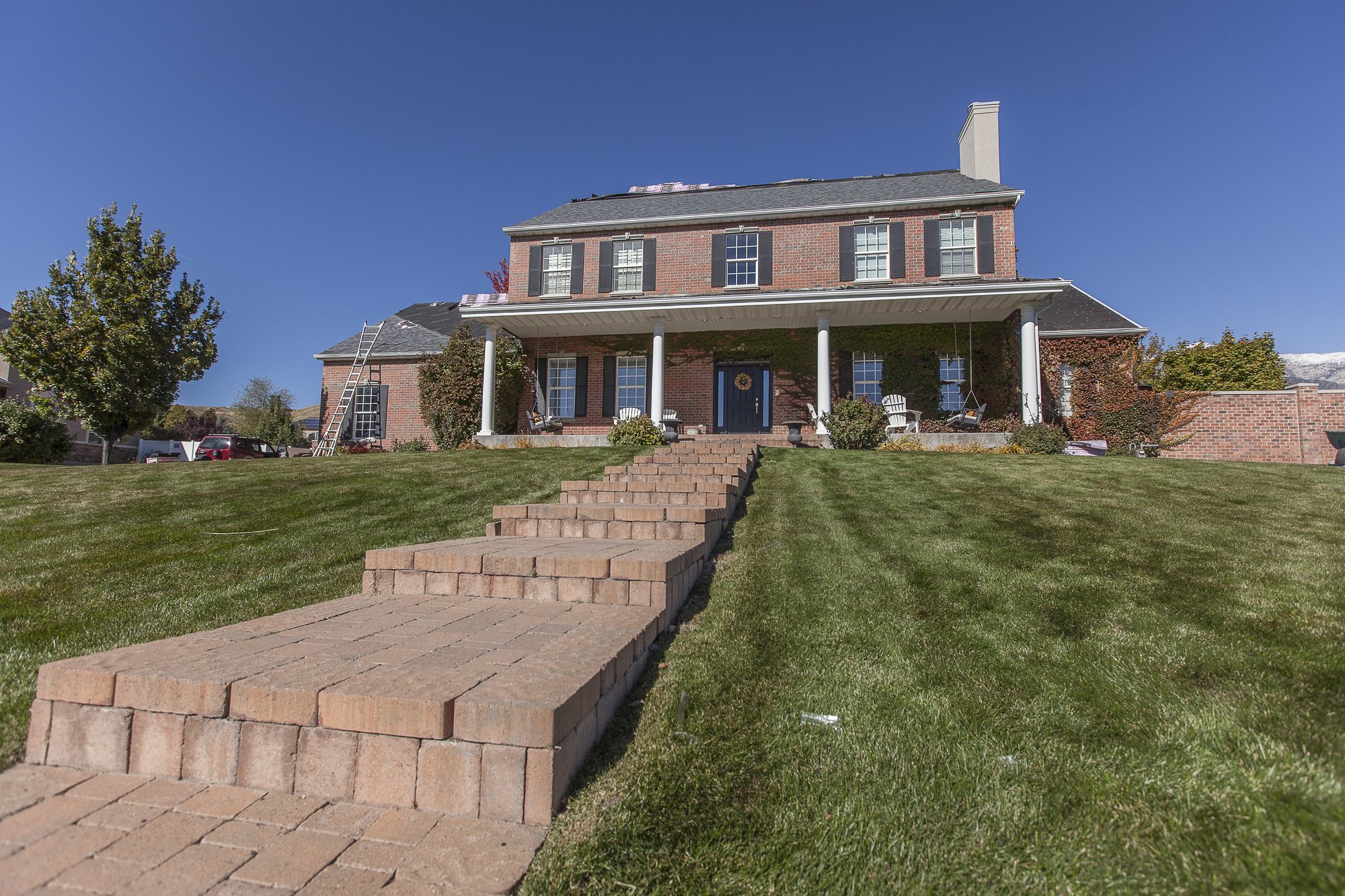 Brick two-story home with columned porch, gray shingle roof partly stripped to underlayment, ladder at side, in Fielding, UT