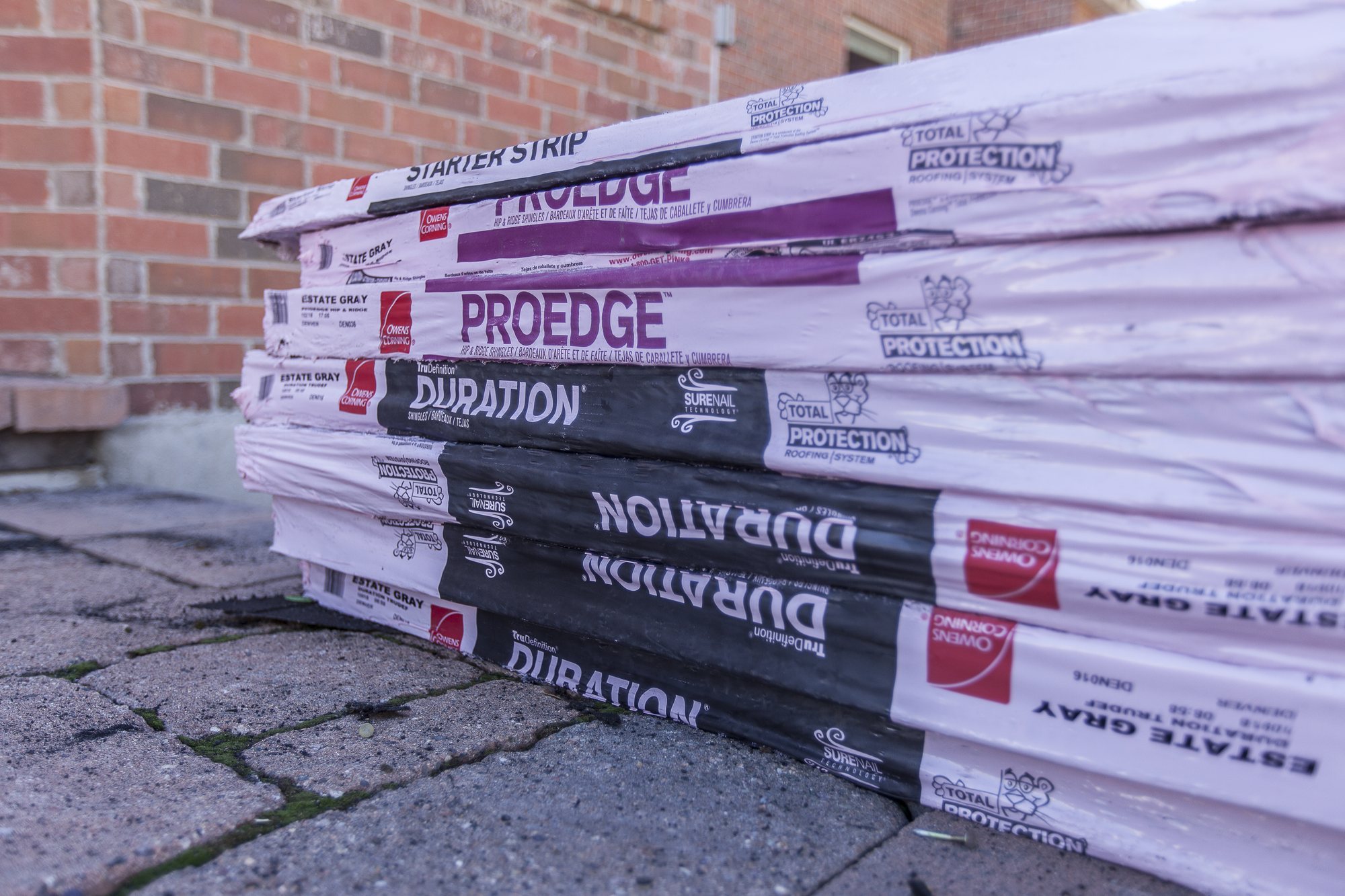 Wrapped Owens Corning Duration and ProEdge shingle bundles stacked on pavers against a brick wall