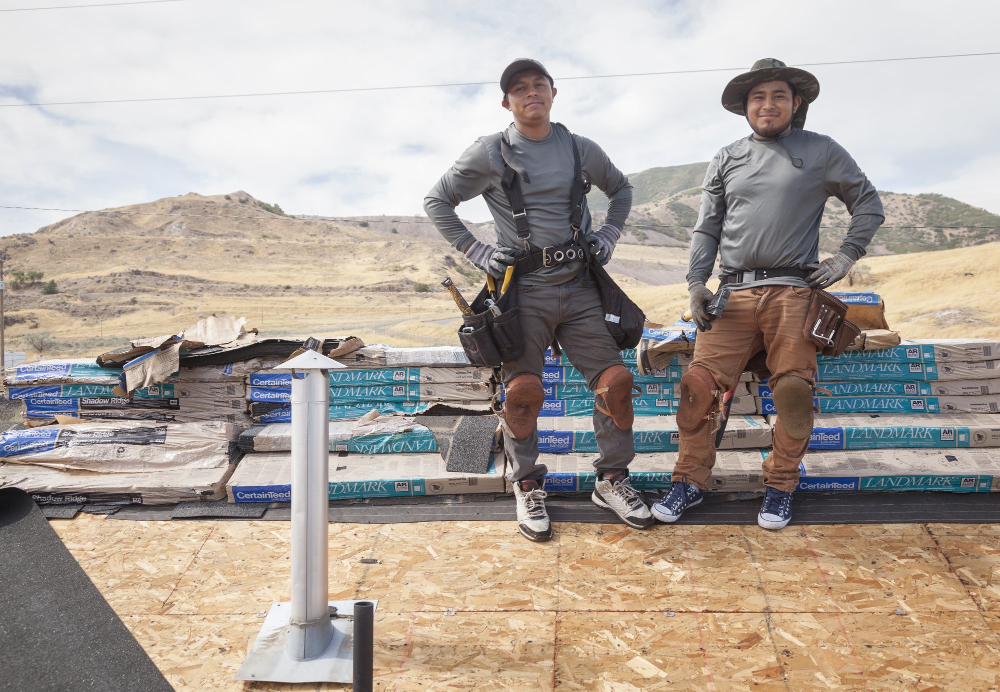 Two roofers in tool belts stand on bare roof decking before stacked wrapped shingle bundles, in West Jordan, UT