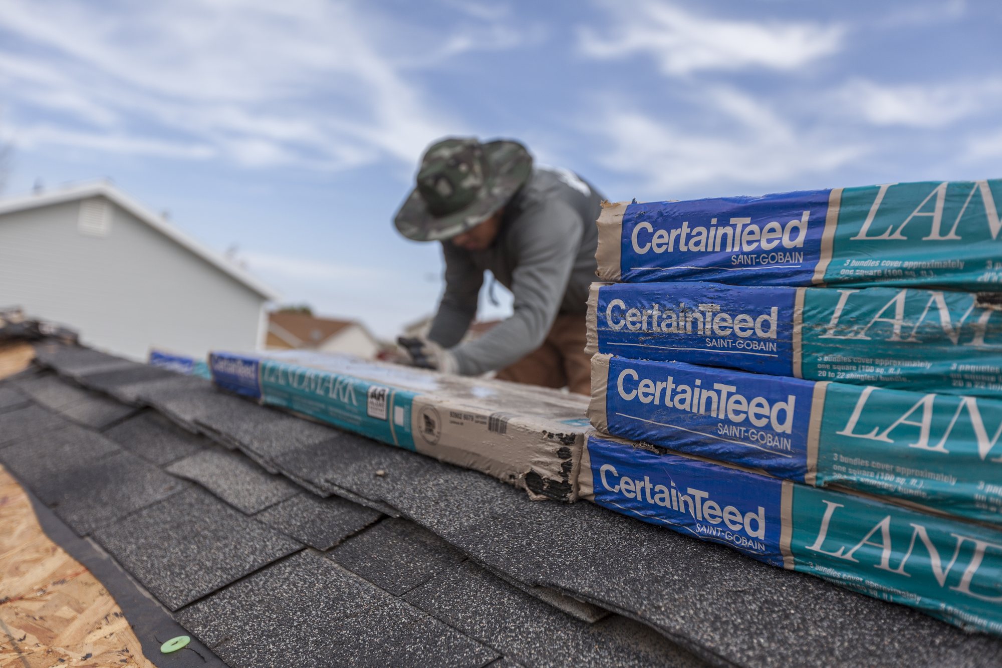 Roofer laying gray asphalt shingles near a ridge next to stacked CertainTeed Landmark shingle bundles