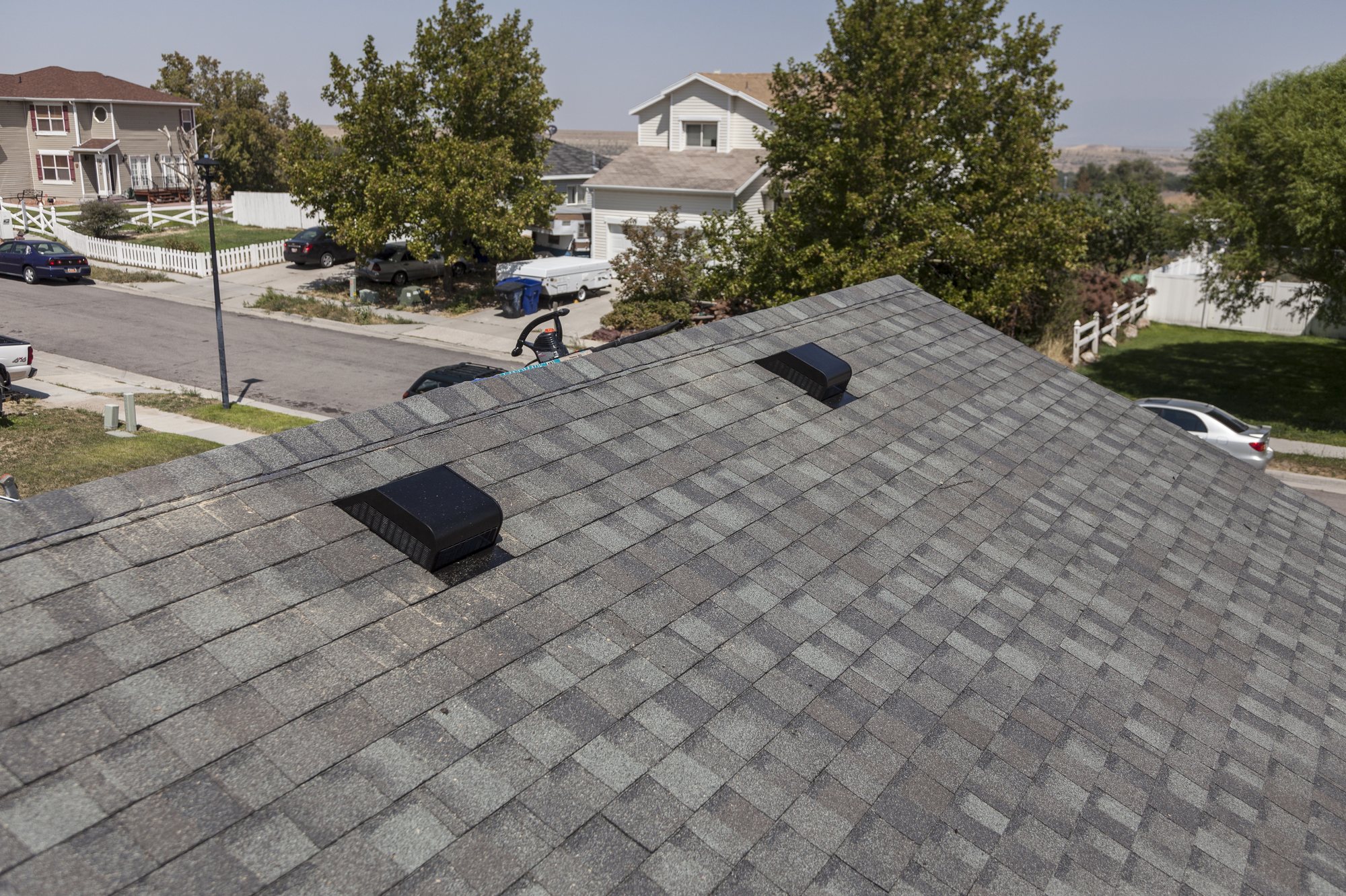 Gray asphalt shingle roof with two black box vents above a suburban street and houses