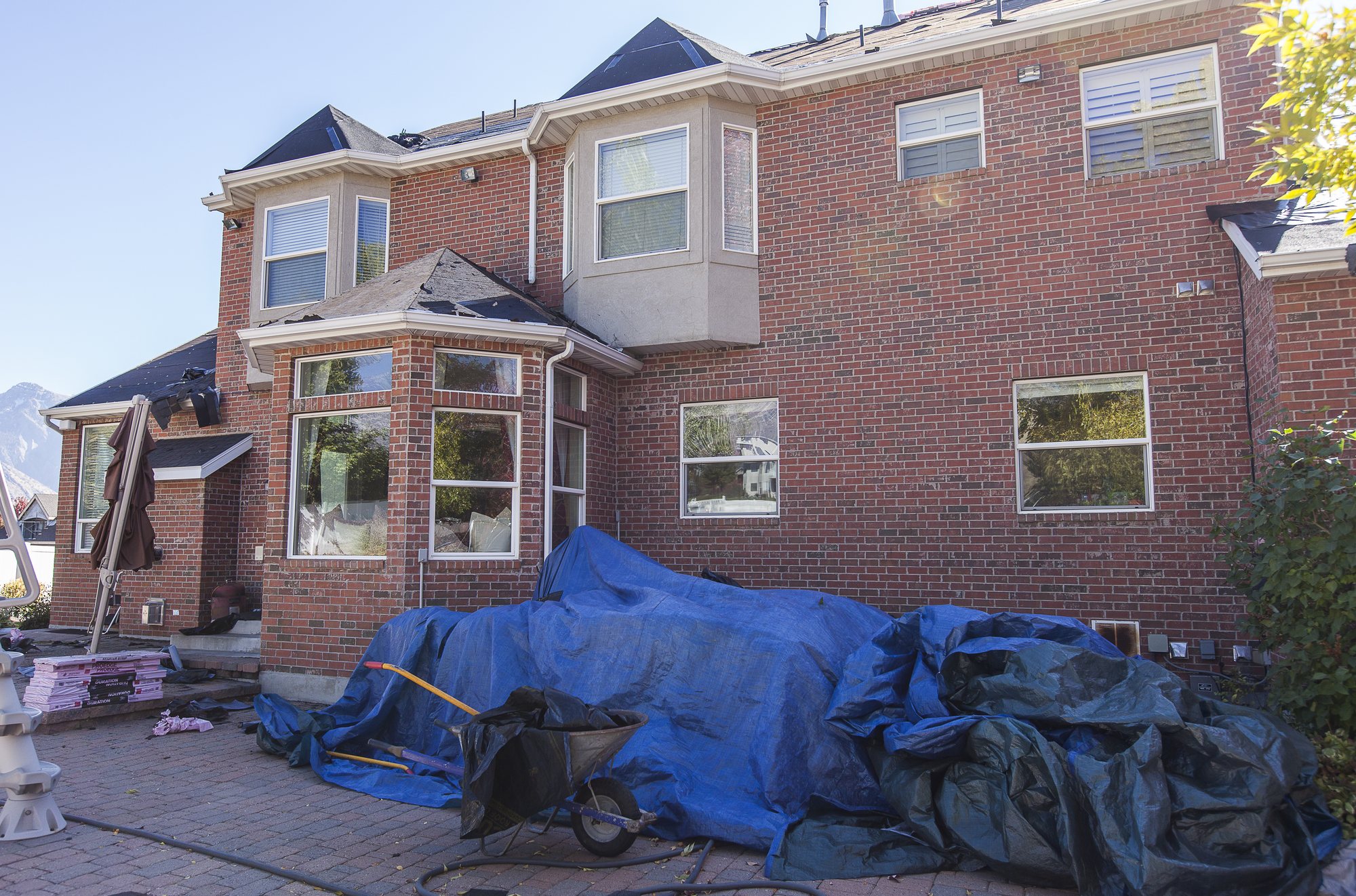 Back of a brick two-story home during a tear-off, with blue tarps spread over the patio and plantings, a wheelbarrow and shingle bundles staged on the pavers, in Clinton, UT