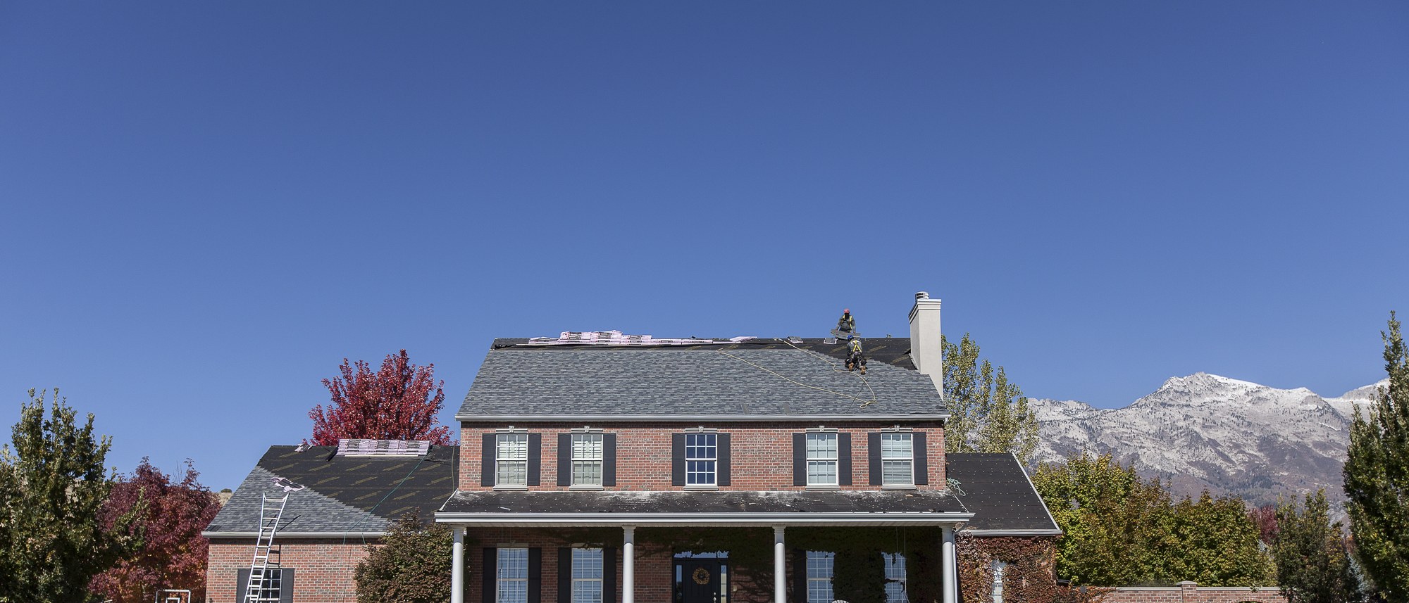 Two harnessed roofers on a gray shingle roof of a brick two-story house, mountains behind