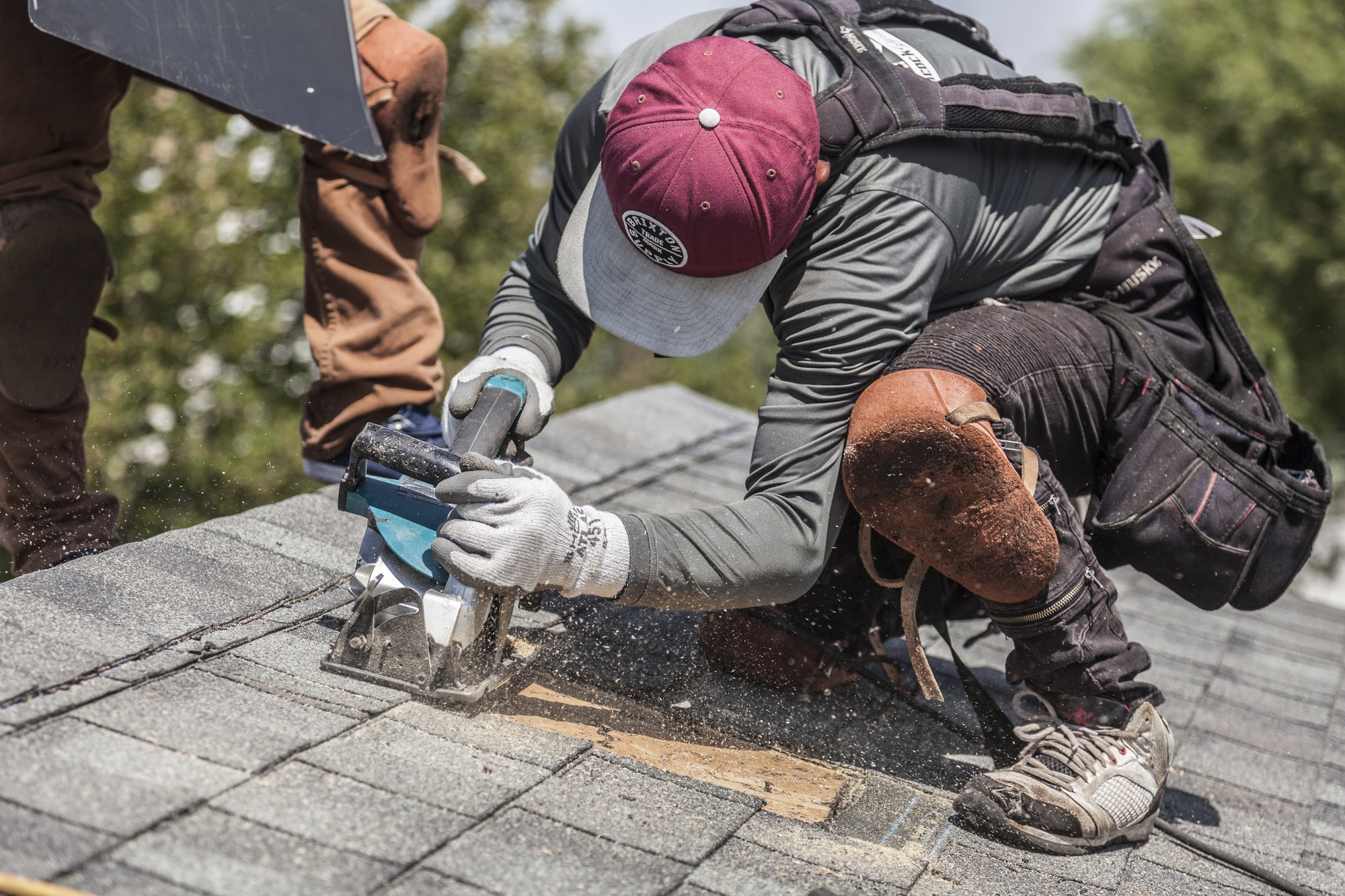Roofer kneeling on gray asphalt shingles cutting with a circular saw, sawdust flying, second worker nearby