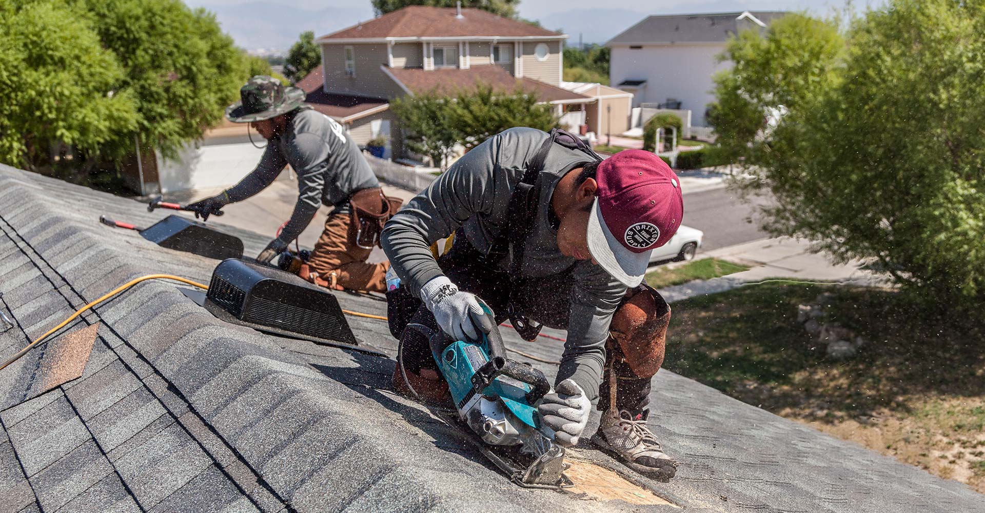 Two roofers on a shingle roof ridge, one cutting decking with a circular saw as sawdust flies