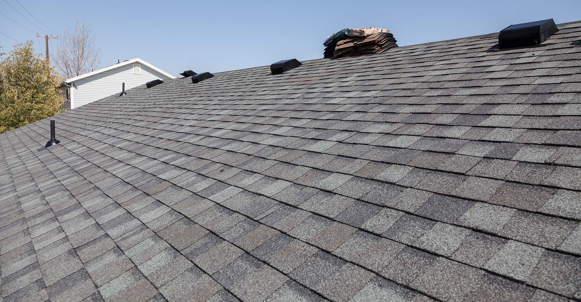 Gray asphalt shingle roof plane with black box vents, a pipe boot, and a shingle bundle near the ridge