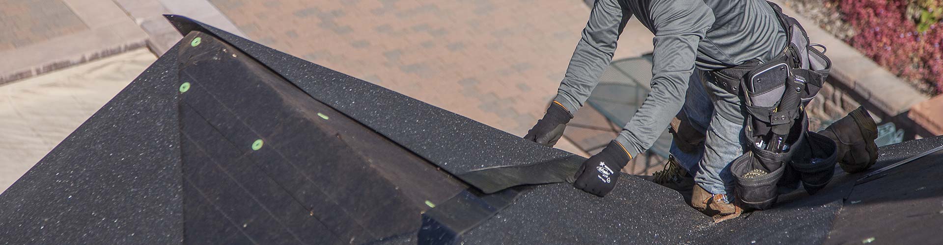 Roofer in gloves and knee pads laying black membrane over a hip, green cap nails visible